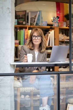 Kendinden emin, orta yaşlı, gözlüklü, stil sahibi bir kadın bir kafedeki masada oturuyor. Yanında duran masada dizüstü bilgisayarı açık ve çalışıyor. Dışarıdan çekilmiş bir fotoğraf. Uzak çalışma. 