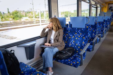 Trenle seyahat ederken dizüstü bilgisayarında çalışan sarışın kadın. Modern bir trende tek başına oturan kendinden emin bir kadın. Uzak çalışma.