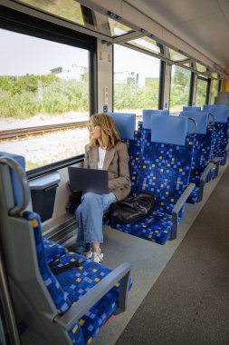 Trenle seyahat ederken dizüstü bilgisayarında çalışan sarışın kadın. Modern bir trende tek başına oturan kendinden emin bir kadın. Uzak çalışma.