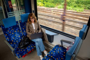 Trenle seyahat ederken dizüstü bilgisayarında çalışan sarışın kadın. Modern bir trende tek başına oturan kendinden emin bir kadın. Uzak çalışma.