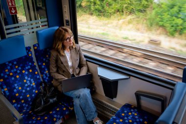 Trenle seyahat ederken dizüstü bilgisayarında çalışan sarışın kadın. Modern bir trende tek başına oturan kendinden emin bir kadın. Uzak çalışma.