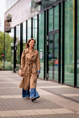 Modern bir ofis binasının önünde yürüyen kumral saçlı, orta yaşlı bir kadının yakın çekimi. Trençkot giyiyor. Tam bacak.