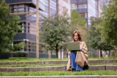 Esmer kadın şehir parkında oturuyor ve iş için dizüstü bilgisayar kullanıyor. Trençkot ve kot giyen kendinden emin orta yaşlı bir kadın. Tam boy çekim. 
