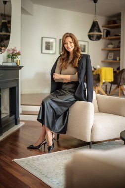 Güzel, gülümseyen orta yaşlı bir kadının koltukta oturup kameraya baktığı bir fotoğraf. Ceket ve etek giyen esmer saçlı kadın. Tam boy çekim.