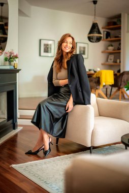 Güzel, gülümseyen orta yaşlı bir kadının koltukta oturup kameraya baktığı bir fotoğraf. Ceket ve etek giyen esmer saçlı kadın. Tam boy çekim.