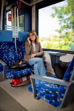 Trenle seyahat eden ve pencerenin kenarında oturan sarı saçlı kadın. Modern bir tren vagonunda oturan ve elinde akıllı bir telefon tutan kendinden emin bir kadın..