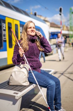 Tren istasyonunda bankta oturan beyaz bastonlu kör kadın portresi. Treni bekleyen ve telefon kullanan görme engelli bir kadın. Günlük kıyafetler. Tam uzunluk. 