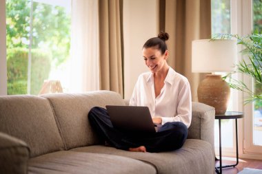 Kendinden emin orta yaşlı bir kadın evden uzakta çalışıyor. Dizüstü bilgisayar kullanırken gülümsüyor. Gündelik görünüş, rahat ve modern iç mekan. Merkez ofis. Serbest çalışma..