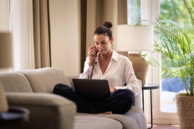 Kendine güveni tam orta yaşlı bir kadın evde uzaktan çalışıyor telefonda konuşurken gülümsüyor ve dizüstü bilgisayar kullanıyor. Sıradan görünüş ve modern iç mekan. Merkez Ofis.