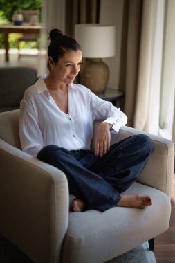 Modern evindeki koltukta rahatlayan esmer bir kadının portresi. Beyaz gömlek, kot pantolon giyen ve hayal kuran beyaz bir kadın. Tam boy çekim. 