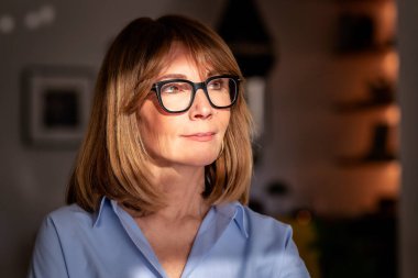 Doğal ışıkta kendinden emin orta yaşlı bir kadının yakın plan portresi. Orta yaşlı, sarı saçlı, kapalı alanda duran ve düşünceli görünen bir kadın. Mavi gömlek ve gözlük takan kendinden emin bir kadın.. 