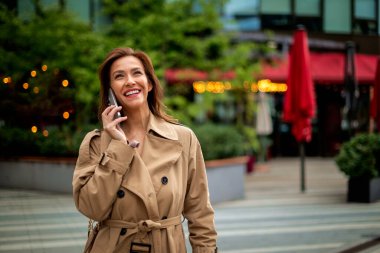 Çekici, orta yaşlı, kumral saçlı bir kadın sonbahar günü şehir caddesinde yürüyor. Kumral kadın trençkot giyiyor ve akıllı telefonunu kullanıyor..