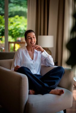 Modern evinde koltukta dinlenen mutlu bir kadın. Beyaz gömlek ve kot pantolon giyen beyaz kadın.. 
