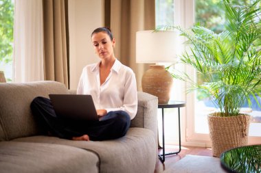 Kendinden emin orta yaşlı bir kadın evden uzakta çalışıyor. Bir dizüstü bilgisayar kullanırken düşünceli görünüyorsun. Gündelik görünüş, rahat ve modern iç mekan. Merkez ofis. Serbest çalışma..