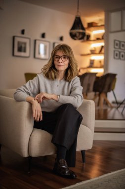 Dairesinde rahat bir koltukta oturan gülümseyen kadının portresi. Gri kazak ve siyah pantolon giyen orta yaşlı bir kadın. Modern iç mekan. Tam uzunluk.