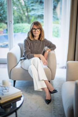 Çekici orta yaşlı kadın evdeki koltukta dinleniyor. Süveter ve pantolon giyen sarışın kadın. Tam boy çekim.