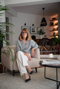 Dairesinde rahat bir koltukta oturan gülümseyen kadının portresi. Gri kazak ve beyaz pantolon giyen orta yaşlı bir kadın. Tam boy çekim.