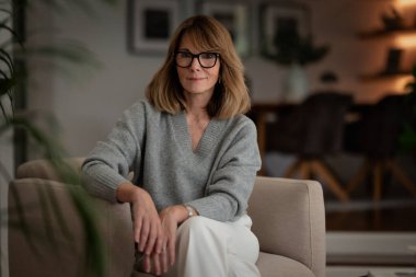 Dairesinde rahat bir koltukta oturan gülümseyen kadının portresi. Gri kazak ve beyaz pantolon giyen orta yaşlı bir kadın..