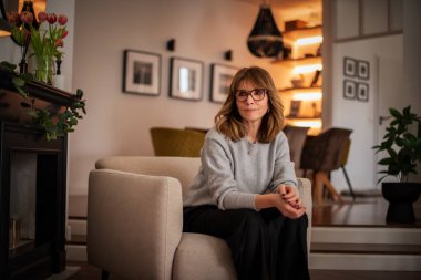 Dairesinde rahat bir koltukta oturan gülümseyen kadının portresi. Gri kazak ve siyah pantolon giyen orta yaşlı bir kadın. Modern iç mimari.