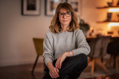 Rahat dairesinde oturan gülümseyen kadının portresi. Gri kazak ve siyah pantolon giyen orta yaşlı bir kadın. Modern iç mimari.