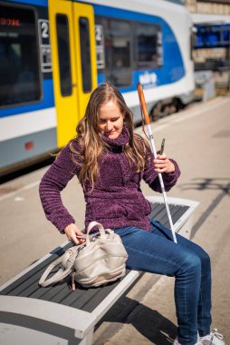 Beyaz bastonlu görme engelli bir kadın tren istasyonunda oturuyor ve birini bekliyor. Tam boy çekim.