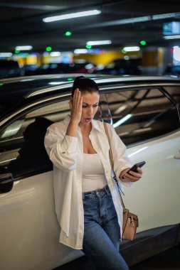 Arabasına yaslanıp telefonunu kullanan güzel bir kadın. Esmer kadın akıllı telefon tutuyor ve hayal kırıklığına uğramış görünüyor..
