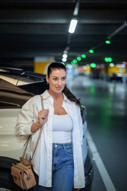 Esmer kadın otoparkta arabasının yanında duruyor.