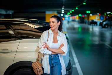 Esmer kadın otoparkta arabasının yanında duruyor.