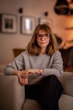 Rahat dairesinde oturan gülümseyen kadının portresi. Gri kazak ve siyah pantolon giyen orta yaşlı bir kadın. Modern iç mimari.