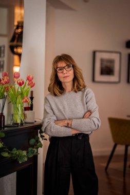 Dairesinde duran gülümseyen bir kadının portresi. Gri kazak ve siyah pantolon giyen orta yaşlı bir kadın. Modern iç mimari.