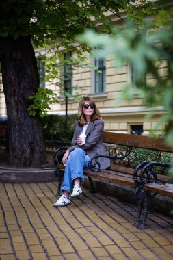 Güneşli bir günde orta yaşlı bir kadın bankta oturuyor. Kendine güvenen kadın spor ceket giyiyor ve güneş gözlüklü kot pantolon giyiyor. Elinde kağıt kahve fincanı vardı. Kahve molası. Tam uzunluk.