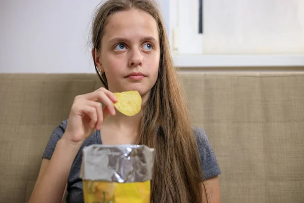 Jeune fille assise sur le canapé et manger des chips du paquet — Photo