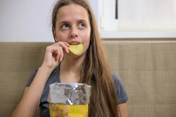 Chica joven sentada en el sofá y comiendo patatas fritas de paquete —  Fotos de Stock
