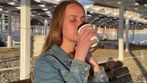 Jovem mulher sentada no banco perto do mar e bebendo café — Vídeo de Stock