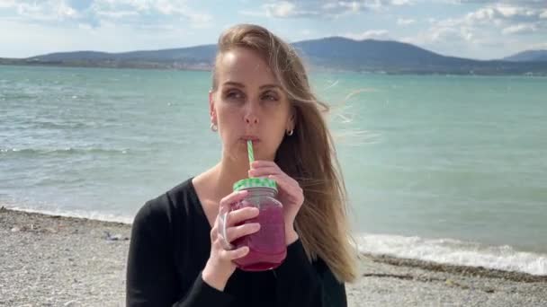 Щаслива жінка п'є ягідну смужку і ходить на узбережжі, повільний рух — стокове відео
