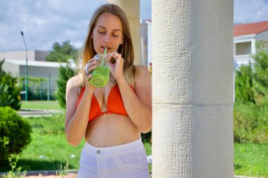 Bikinili sarışın kadın tatil köyünde yeşil smoothie içiyor. 