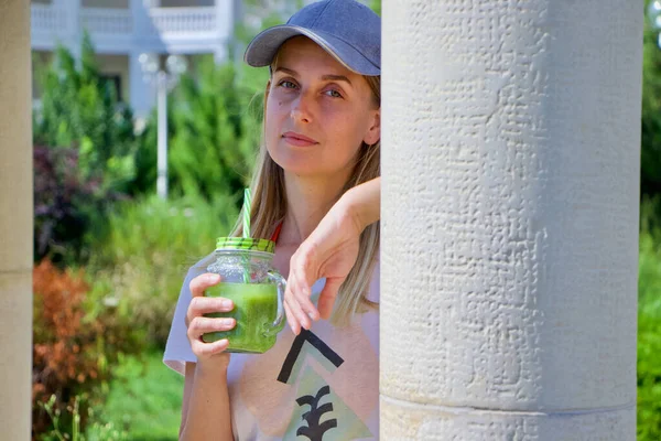 Woman in cap holding glass of green smoothie and drinking it — Stock Photo, Image