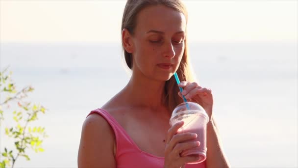 Veganerin im rosafarbenen Kleid entspannt an der Küste mit Beeren-Smoothie — Stockvideo
