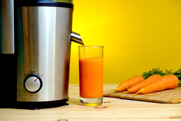 Juicer and carrot juice in glass on yellow background