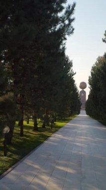 Özbekistan 'ın Taşkent kentindeki huzurlu bir parkta yüksek çam ağaçları tarafından çerçevelenmiş Dünya Anıtı manzarası. Güneş ışığı, yürüyüş yolunu hafifçe aydınlatır, doğa, kültür ve kentsel güzelliği ön plana çıkaran sakin bir atmosfer yaratır..