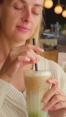 Beyaz örgü süveter giyen sarışın bir kadın sıcak bir kafede pipetle buzlu kibrit latte içiyor. Yeşil ve kremsi tonlu katmanlı içecek, sakin ve rahatlatıcı bir anı yansıtıyor, modern bir yaşam tarzını ve iç mekandaki huzuru yansıtıyor..
