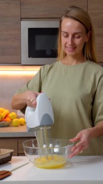 Kadın modern bir ev mutfağında malzemeler hazırlıyor, elektrikli el mikseri kullanarak cam bir kasede yumurta çırpıyor ve gülümsüyor ve yemek pişirme sürecinin tadını çıkarıyor..