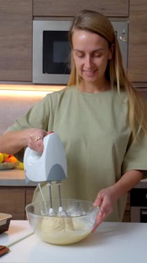 Gülümseyen kadın modern bir mutfakta elektrikli el mikseri ve cam kase kullanarak ev yapımı hamur hazırlıyor. Tezgahta taze meyveler ve yakınlarda pişirme malzemeleri var..