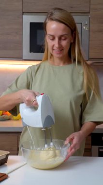 Ev mutfağındaki cam kasede kek hamuru karıştırmak için elektrikli el mikseri kullanan gülümseyen kadın. Pişirme süreci, ev yapımı tatlı hazırlama, günlük yemek pişirme, rahat yaşam tarzı ve yemek rutini.