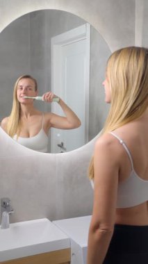 Banyodaki aynaya bakarken elektrikli diş fırçasıyla dişlerini fırçalayan genç bir kadın. Modern minimalist iç mekanlarda günlük hijyen rutini.