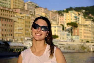 Sahilde güneş gözlüğü takan genç bir model. İtalya, Liguria, Camogli plajı. Arka planda renkli evler var.