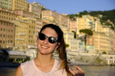 Sahilde güneş gözlüğü takan genç bir model. İtalya, Liguria, Camogli plajı. Arka planda renkli evler var.