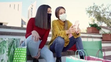 Yeni alışveriş alışkanlıkları: iki güzel beyaz kadın bir meydanın basamaklarında, koruma maskesi takan birçok renkli alışveriş torbası arasında oturuyorlar. İnternette farklı fiyatları aramak için akıllı telefon kullanıyorlar.