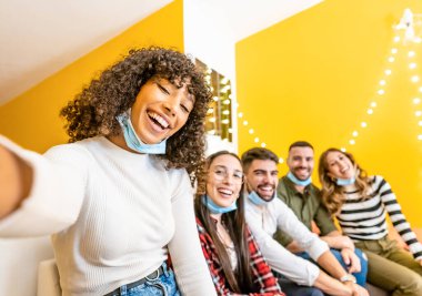 Teknolojik eğlence evinde eğlenen çok ırklı genç bir grup güzel siyah İspanyol kadın, Coronavirus maskesi takmış arkadaşlarına akıllı telefonuyla fotoğraf çekiyor.