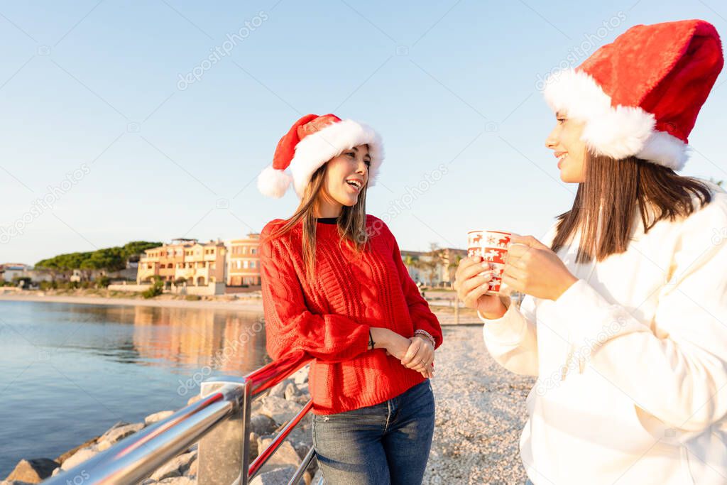 Beautiful young caucasian women standing outdoor talking with a Christmas mug in hands wearing Santa hat - Sunset sea winter vacation for two female friends at Golfo Aranci, Sardinia